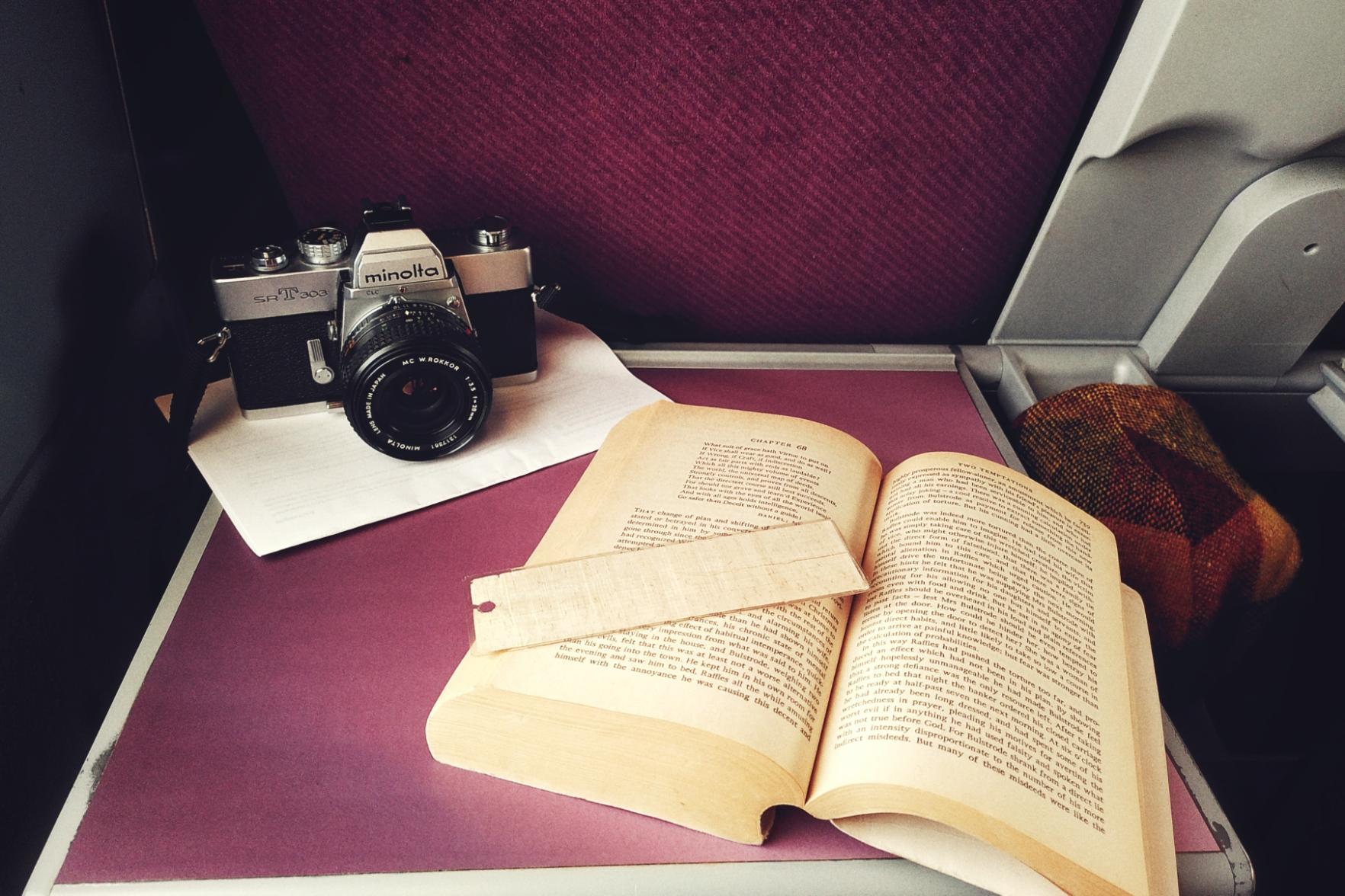 A horizontal colour photograph of a purple train table with a film camera and an open book on it. The film camera is resting on some folded paper. Next to the table is a yellow and red flat cap hanging on a water holder extrusion.