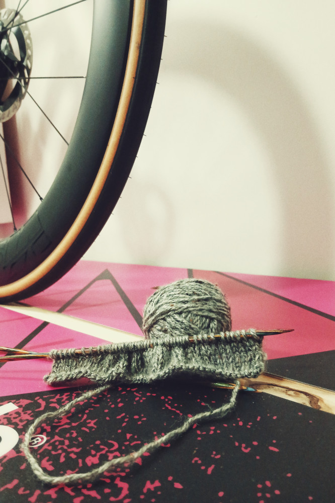 A vertical colour photograph of a small strip of grey knitting on wooden needle rests on a pink plinth by a bicycle wheel.