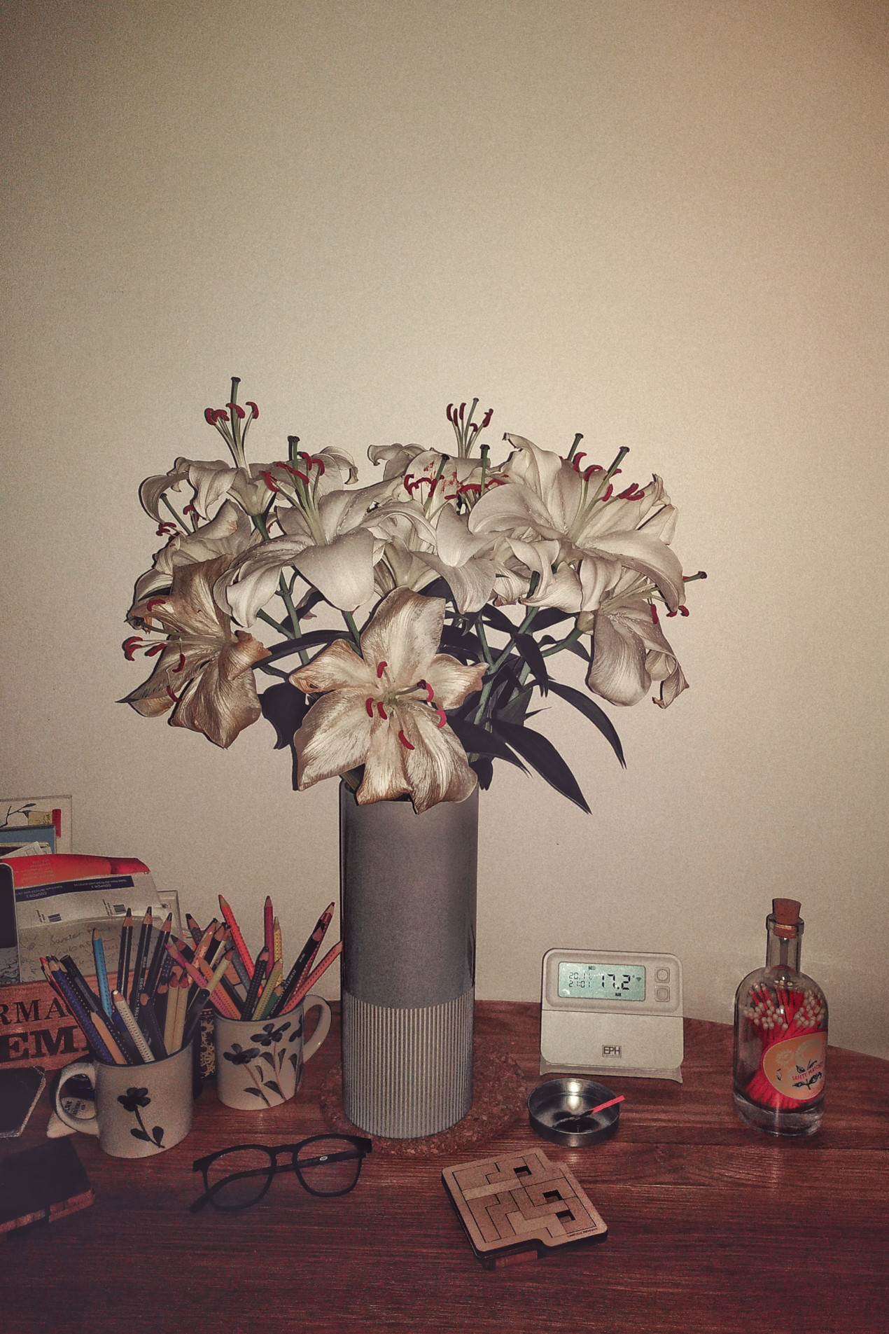 A vertical photograph of some fading white lilies in a grey vase on a wooden table. Around the vase are some art supplies in cups, a thermostat, a pair of glasses, some long matches in a glass jar.