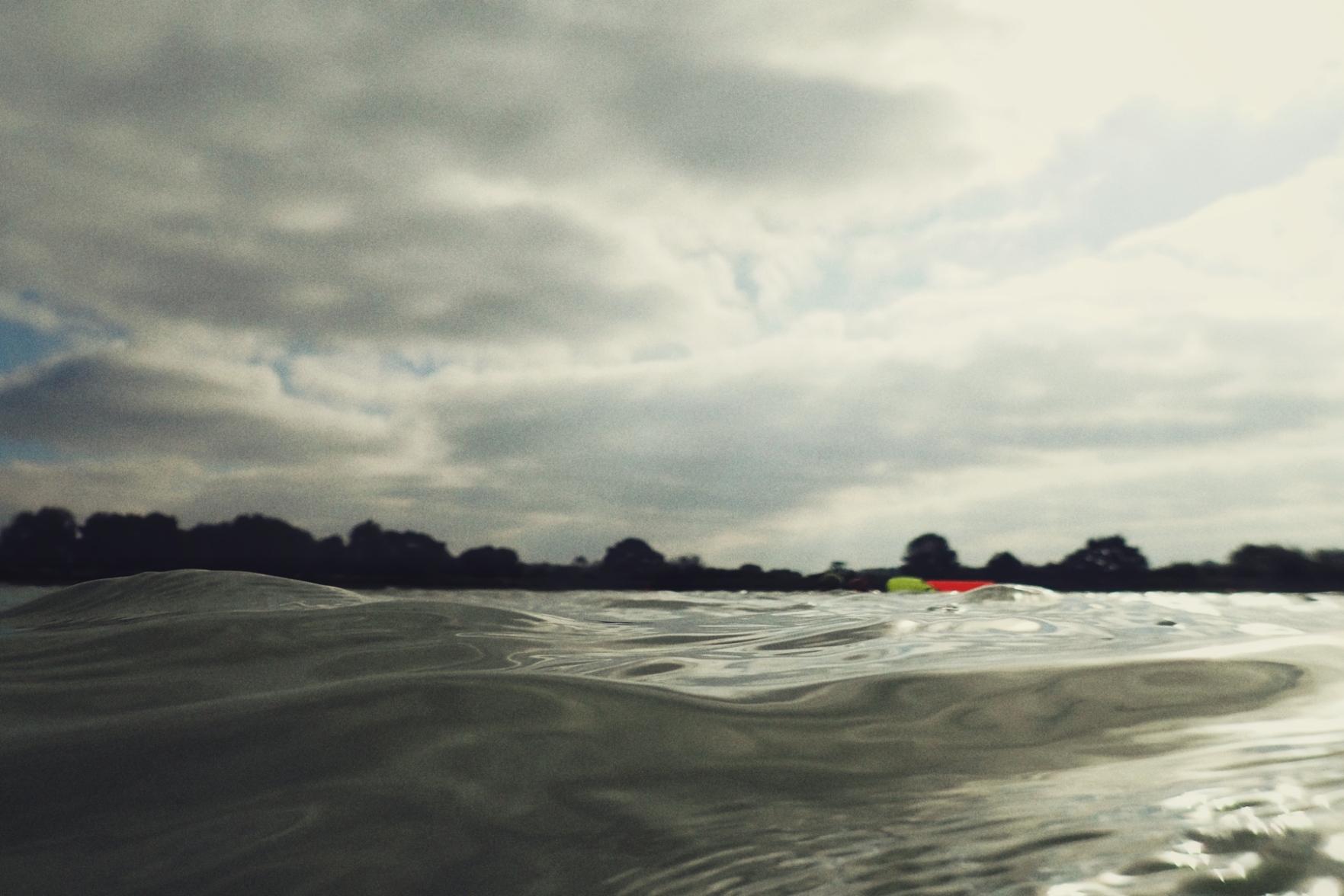 A horizontal colour photograph of a lake from the water. The water is making repeated rounded bumps. In the distance are two spots or bright colours where people are swimming. Further on is a line of trees. The sky is cloudy with some bits of blue.