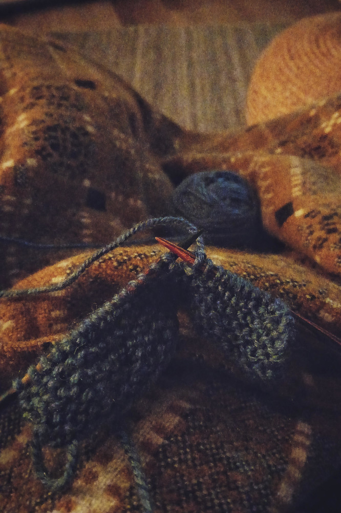 A vertical colour photograph of a small knitted work on wooden needles. The yarn is blue. The ball rests in my legs. My legs are covered by a yellow and grey wool blanket.