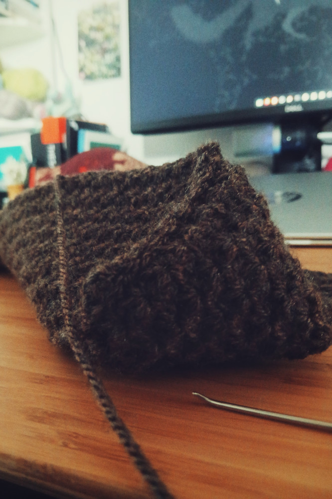 A vertical colour photograph of a brown crochet pouch in the progress of being stitched together. The pouch is on a desk with a background of desk paraphernalia just visible. The pouch slouches as one corner is stitched but not the rest.