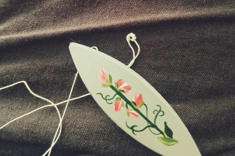 A horizontal colour photograph of a blue tatting shuttle with a painted sweet pea on it. Above is a tiny round of tatting work in a white thread.