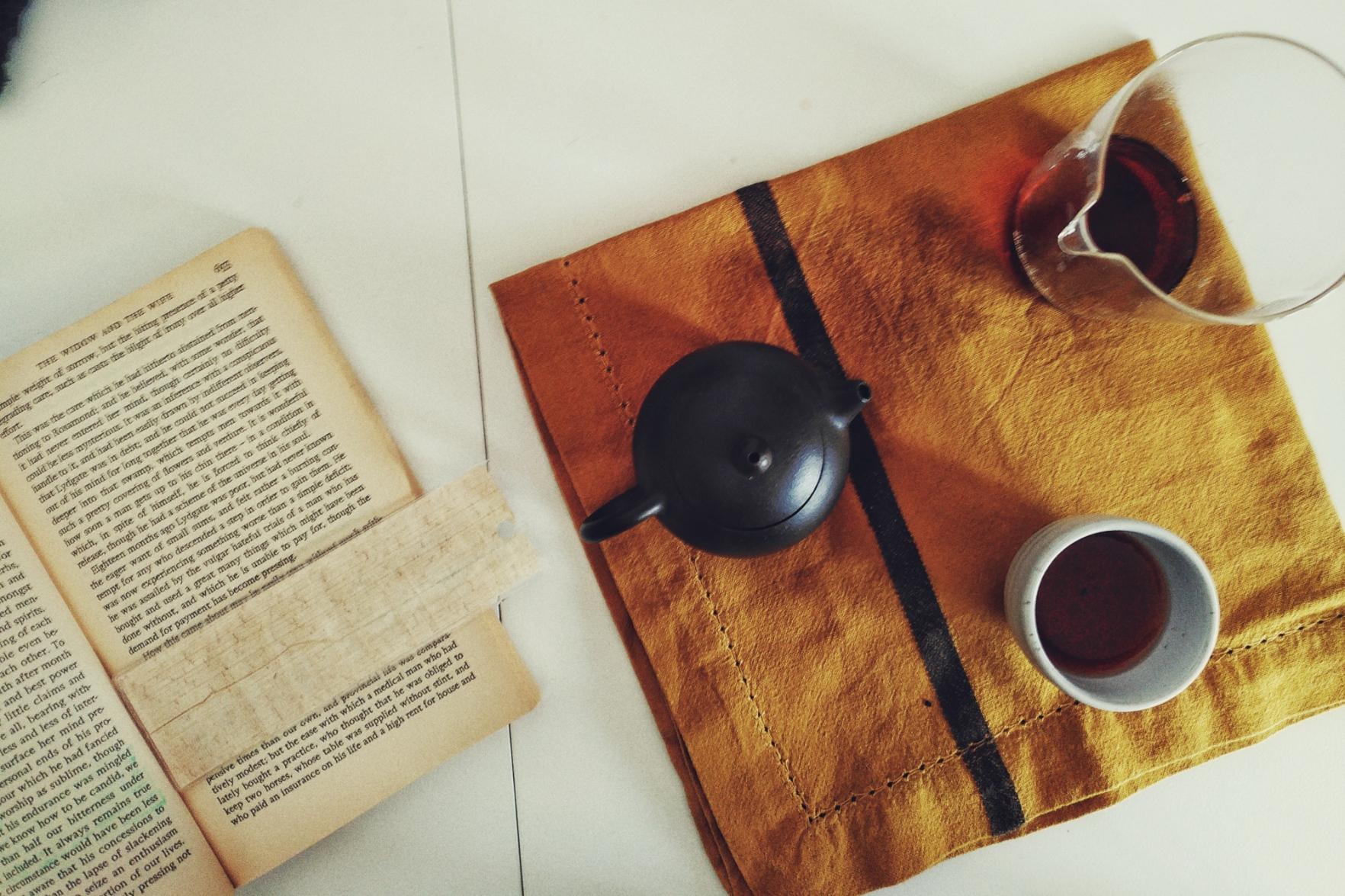A horizontal colour photograph of a white table with a tea set on a mustard coloured cloth. There is a small brown teapot, a fairness jug with some tea in it, and a small white cup with a little bit of dark tea left in it. To the left of the cloth is a open paperback book with a papyrus bookmark marking the current reading line.