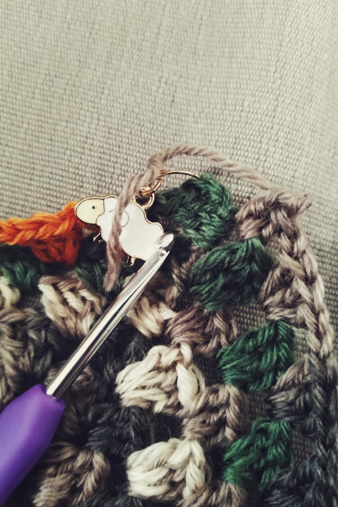A vertical colour photograph of a corner of a multi coloured granny square. One side has a stitch marker in the shape of a sheep. The other side has two stitches and a small piece of yarn left to work with. It's not big enough to complete the last couple of stitches. A metal crochet hook rests on the work.