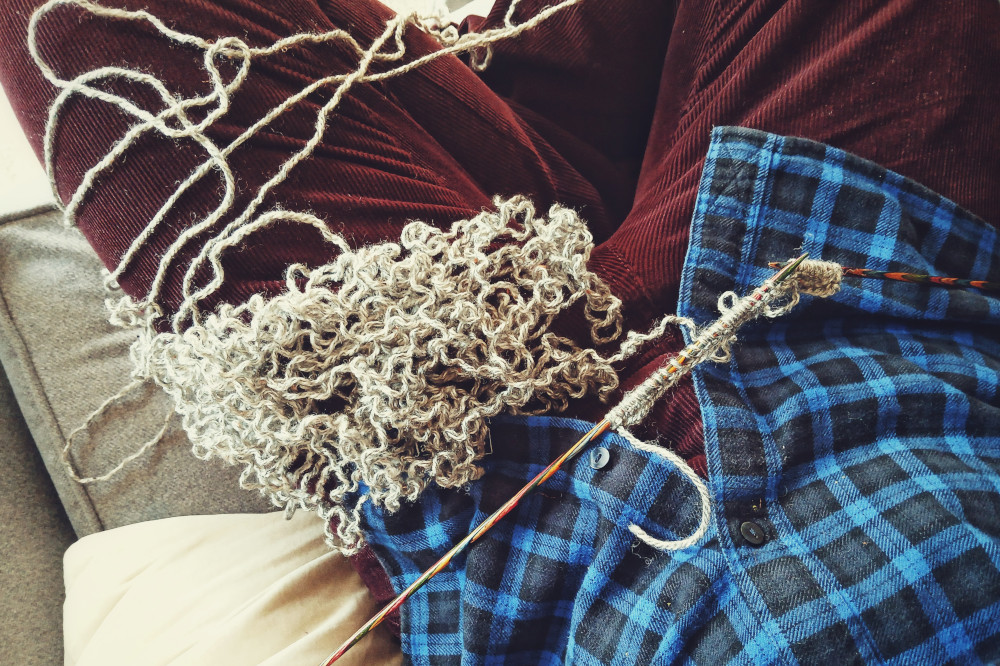 A horizontal colour photograph of a pile of grey yarn curled up after having been fogged from the needles. The yarn rests on my crossed legs. I'm wearing a burgundy corduroy and checked dark blue and black shirt.