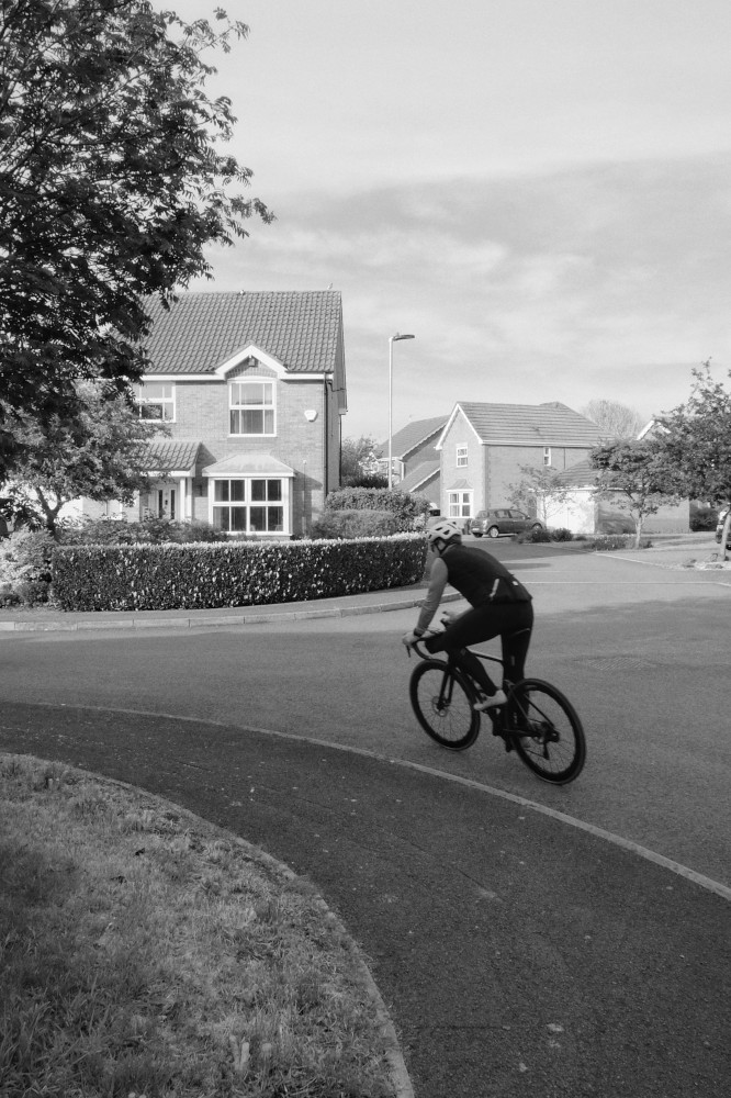 A vertical black and white photograph of a cyclist on a road going around a curved pavement curb with a tree at the left edge corner of the frame.