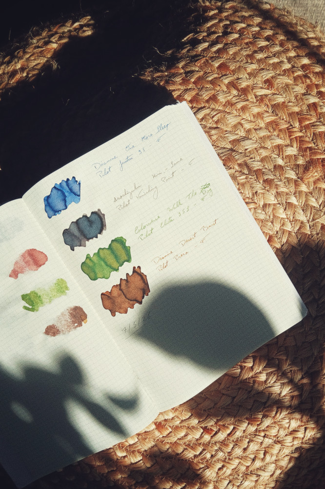 A vertical colour photograph of ink swatches on a notebook with a grid pattern on the paper. The sun falls across the paper with a diagonal streak of shadows on the bottom of the page. The opposite page is visible with the back of previous swatches seeping through. The inks are blue, grey, green, and brown.