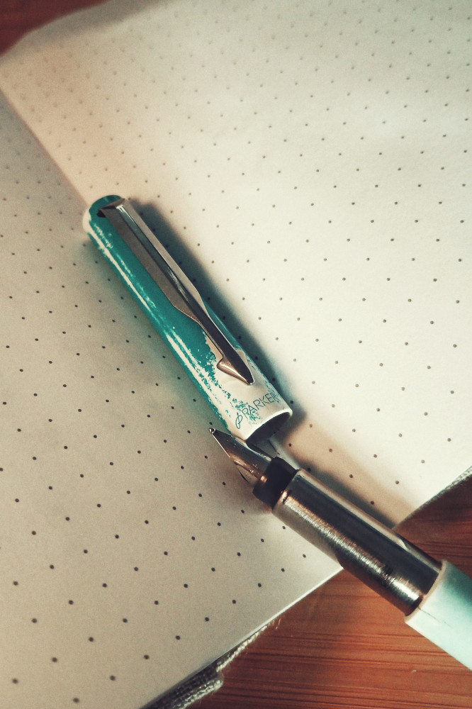 A vertical colour photograph of a fountain pen with the cap open on a dotted notebook. The nib and grip are silver looking. The cap has seen better days, the colour gone in some spots with small cracks at the bottom edge.