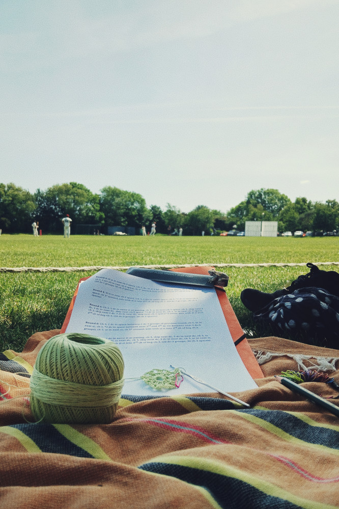 A vertical colour photograph of a small light green crochet work. It rests on a paper where instructions are written. The ball of thread is on an orange, black, and yellow blanket. Above, in the upper half of the frame is a cricket team un white playing under a blue sky.