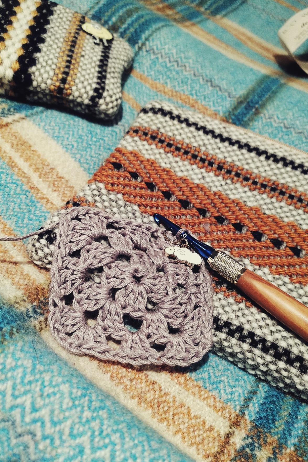 A vertical colour photograph of a lavender granny square with a crochet hook still attached to it. By the crochet hook is a stitch marker in the shape of a sheep. The work rests on a cream, orange, and black woven purse. A other smaller woven purse in yellow, black, and cream is visible at the top left of the image. The image is set on a background of a blue and yellow checkered blanket.
