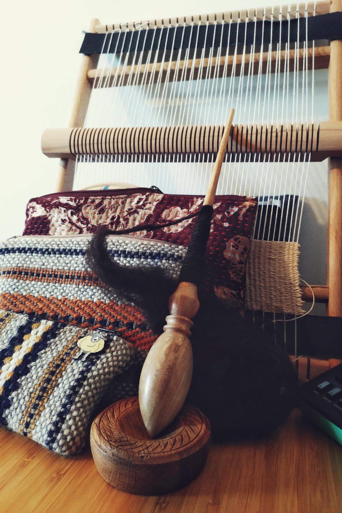 A vertical colour photograph of a wooden Russian style spindle with some dark brown wool spun on it. The spindle rests on a wooden base. Next to it and behind it are three pouches. Two are woven and mostly cream.and grey with colour accents, one is red fabric with a thread all over it. Supporting all this is a frame loom put upright against a white wall.