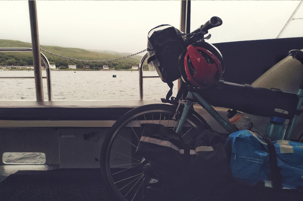 A horizontal colour photograph of the front of a bicycle on a ferry. In front of the bicycle are panniers and bags. Behind is the sea and land shrouded in clouds.