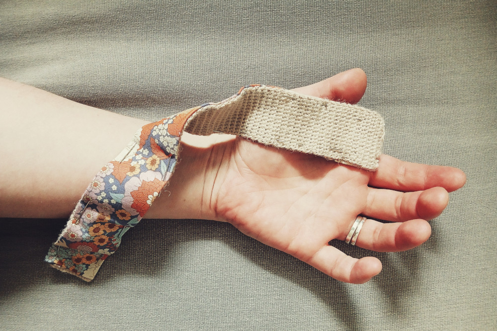 A horizontal colour photograph of a hand and forearm resting on a grey sofa fabric seat. A strip of fabric and crochet held together by pins is twisted across my hand and wrist to show both side of the work.