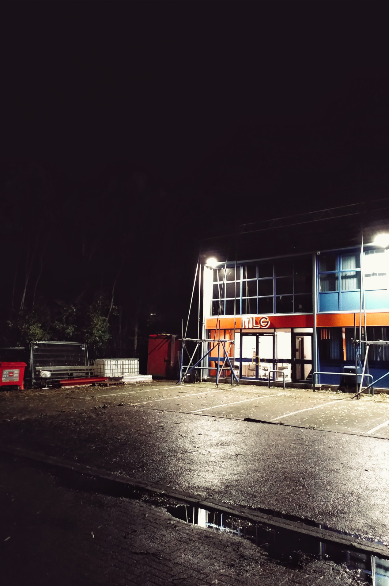 A vertical photograph of part of a warehouse at night with scaffolding in front and blind lights creating deep shadows around. In the foreground is a dark puddle reflecting the light. In the background we can guess at foliage behind bins and fencing materials.