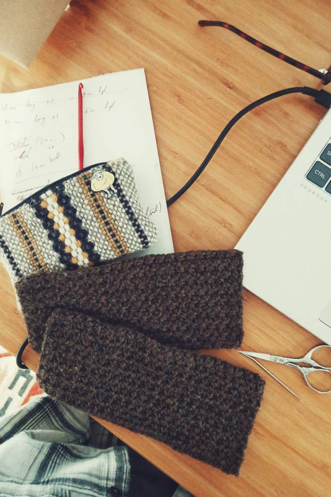A horizontal colour photograph of two crochet brown rectangles on a desk. They rest on a notepad with a small woven pouch above them. On the right hand side the corner of a silver coloured laptop is visible.
