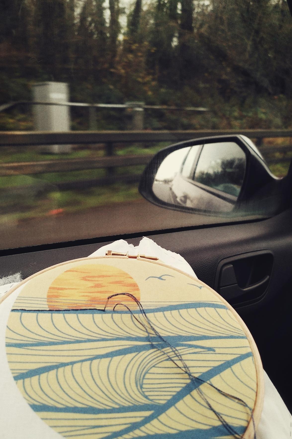 A vertical colour photograph of an embroidery loop on a car. The embroidery is a stylised sunset printed on cotton to guide the stitching. The hoop rests by the passenger window. Outside, vegetation and some barriers are blurred from the speed of the car.