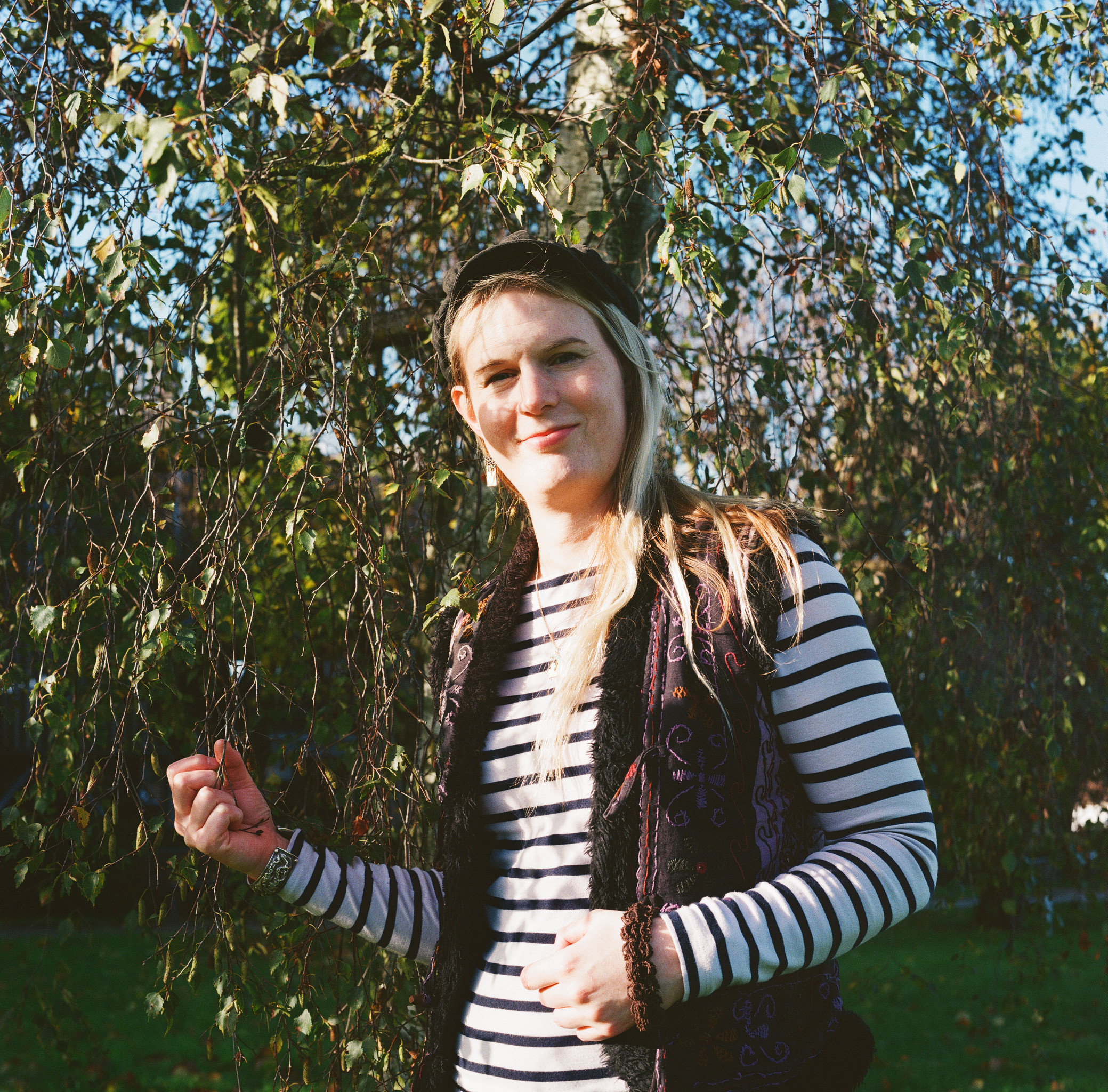 A colour photograph of a woman from the waist up. She is standing in the midst of branches, holding a dangling brand with one hand. The other hand rest by her stomach. She has long blond hair and is wearing a cap.