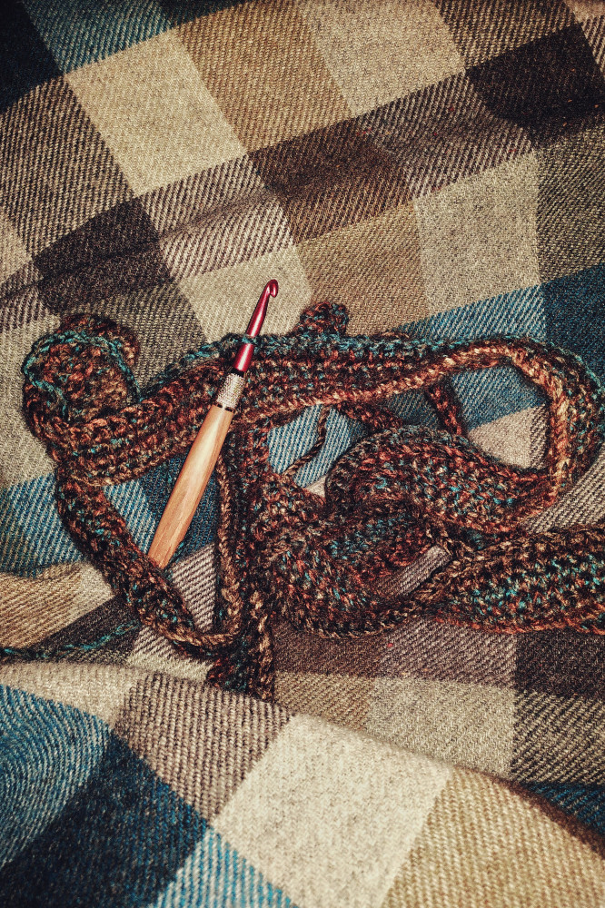 A vertical colour photograph of a crochet scarf in progress. It is in a brown and blue yarn. It is curled on a wollen blanket with grey, blue, and green colours. A crochet hook is attached to the scarf.