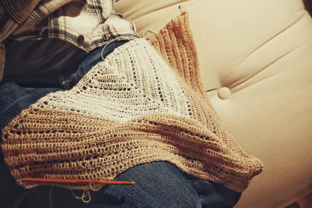 A horizontal colour photograph of a crochet shawl in progress. It drapes across my bent legs in blue jeans. The top is in a white yarn whilst the bottom is in a creamish pink yarn.