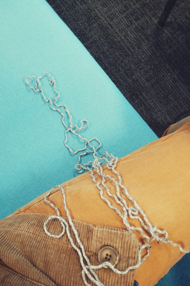 A vertical colour photograph of a grey twisty yarn that has just been frogged on a blue sofa seat. My legs are visible in mustard coloured jeans at the right hand side of the image.