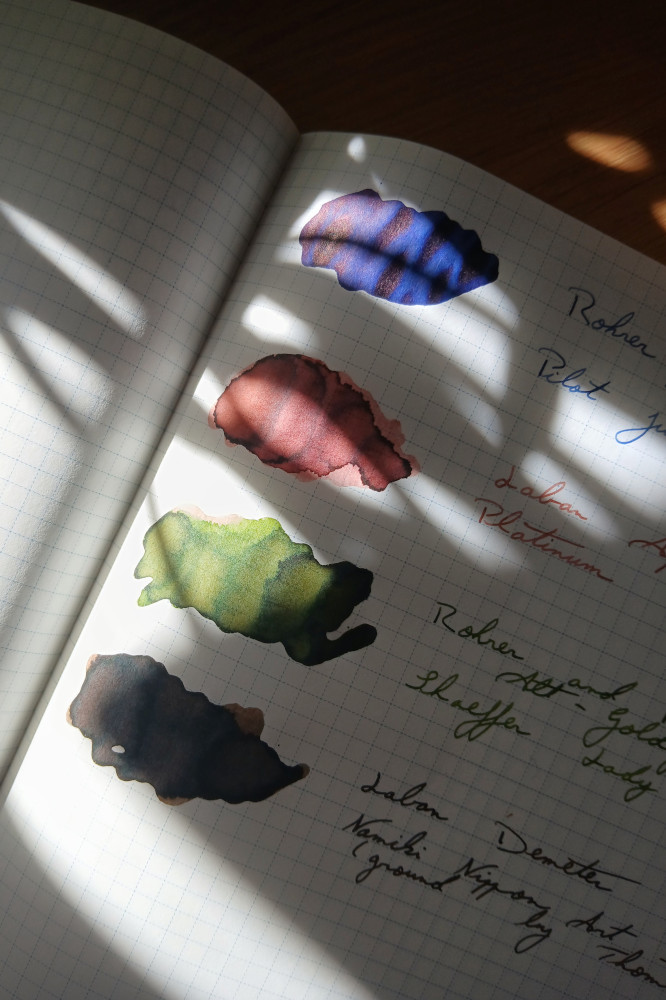A vertical colour photograph of ink swatches on a notebook with a grid pattern on the paper. The sun is dappled and falls in pools across the paper. The inks are blue, pink, green, and brown.