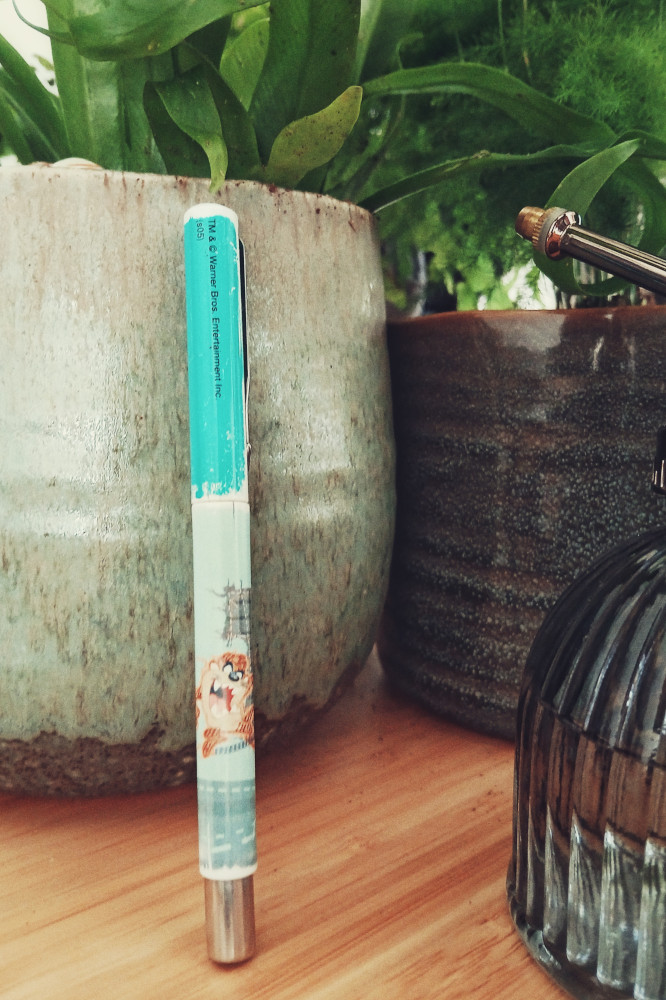 A vertical colour photograph of a teal and light green fountain pen standing upright by a grey plant pot. The greenery of the potted ferns is visible at the top of the image. In the foreground, cut by the frame is a vaporiser.