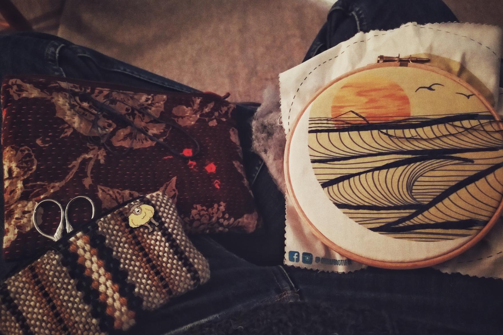 A horizontal colour photograph of an embroidery work on my laps. My legs are crossed legged and in blue jeans. The embroidery hoop is on the right with a stylised sunset printed over the cloth. On the left leg are a couple pouches. One is a deep burgundy with flowers and stitches visible on the top. The other is woven with cream, yellow, and black wool, and had a pin sheep on a corner. Small scissors handle poke out of it.