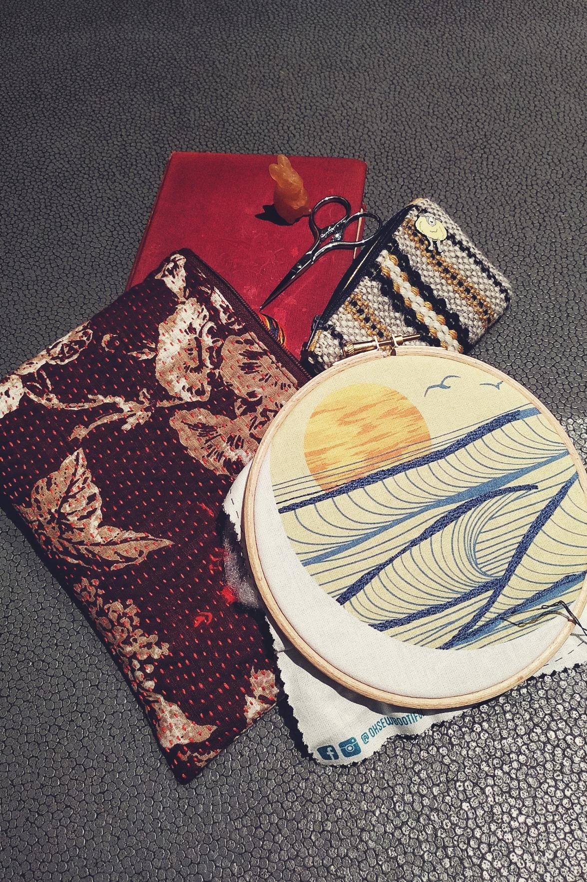 A vertical colour photograph of an embroidery loop resting over two pouches and one red book. On the book is amber rabbit and a small pair of scissors. One of the pouch is a deep Bordeaux and made of fabric. The other one is smaller and woven with cream, grey, and yellow wool. The embroidery in the hoop is on a pre-printed fabric and represents a stylised sunset over waves. Some of the top of the waves have been embroidered with a dark blue thread.
