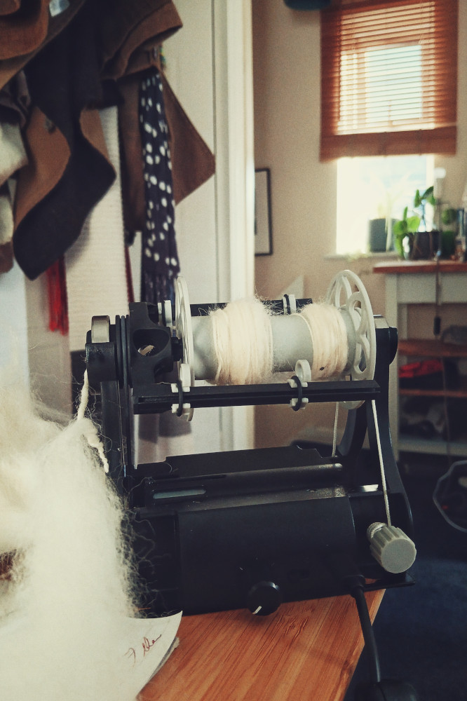 A vertical colour photograph of a small black e-spinner with white floof spun on it and unspun next to it on a wooden desk. In the background is a door with coats and scarfs draping down, a stair landing with a table, and behind a window with wooden blind halfway down.