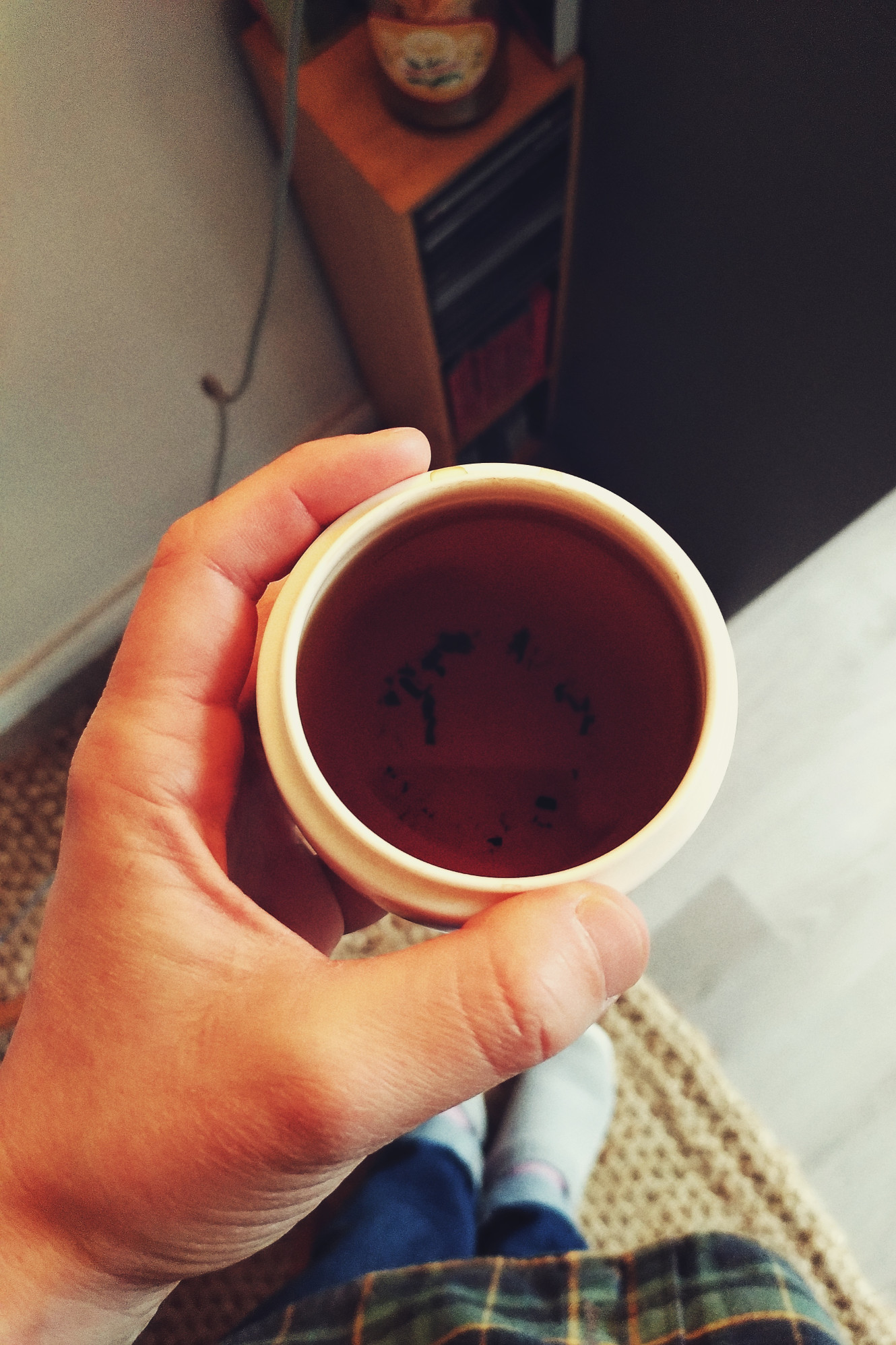 A vertical colour photograph of a cup of dark tea held in the hand over a half carpeted floor, half hard floor. In the background there is a small CD tower.