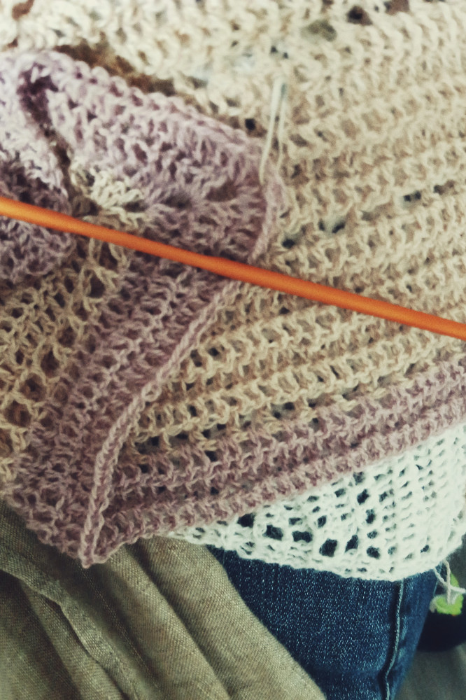 A vertical colour photograph of a section of a crochet shawl with a fingering weight yarn. The colours are white, cream, and violet pink. An orange crochet hook goes through the work. The work rests on my legs. I am wearing blue jeans, just visible at the bottom right of the image.