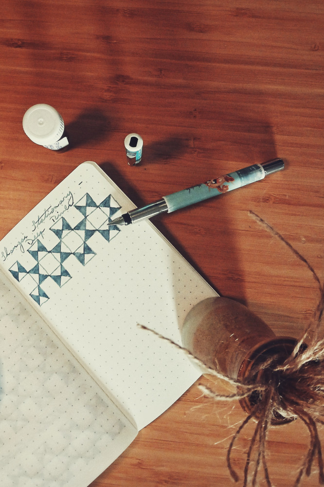 A vertical top down colour photograph of a fountain pen resting sideways by a small dotted notebook with a swatch of a blue grey ink. The ink sample is in a vial above the notebook. Next to it is the pen cap, set upright on the wooden desk. A small ceramic vase with twine in it holds the notebook down.