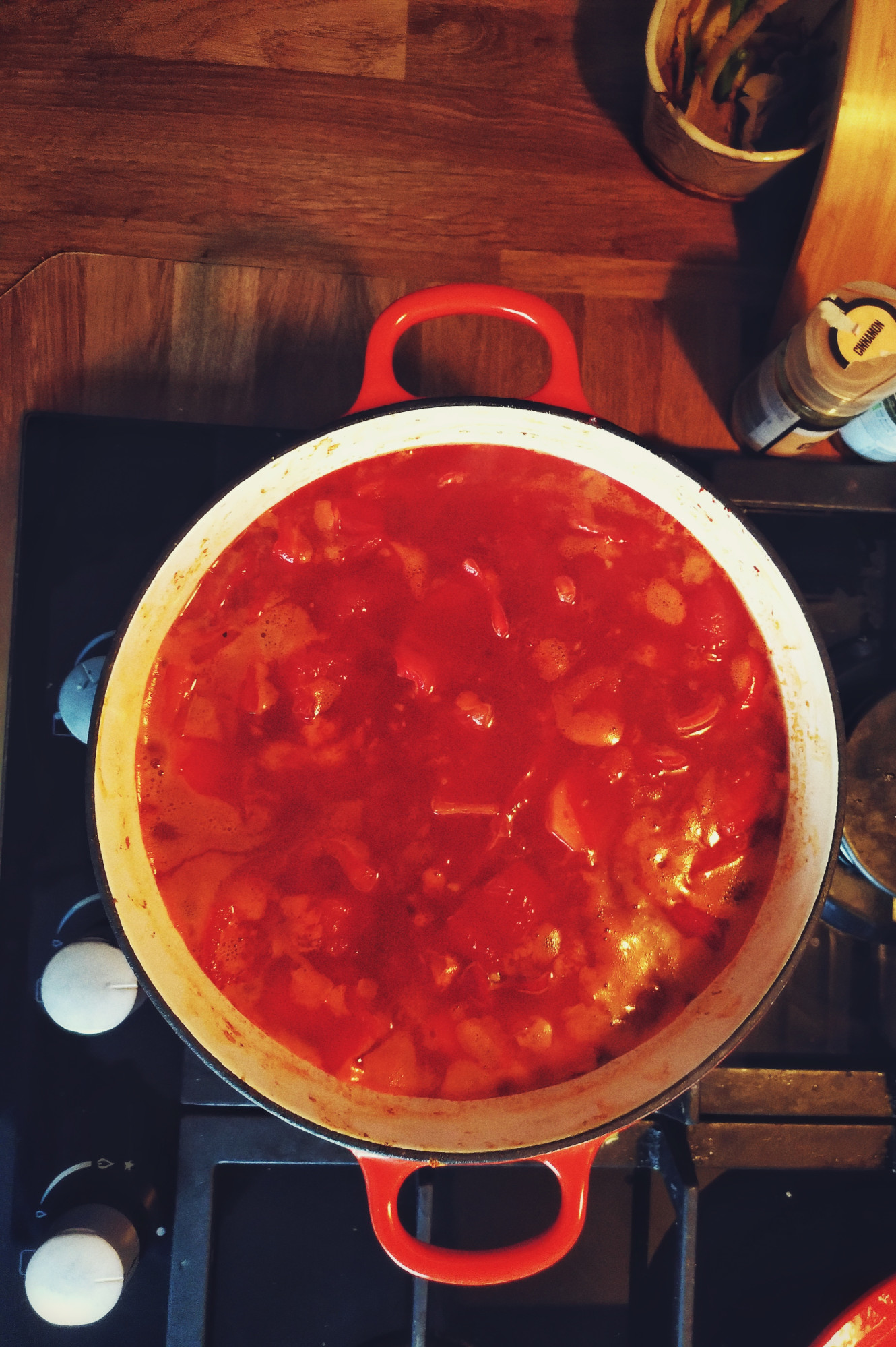 A vertical colour photograph of a red soup with chunky vegs cooking in stock in a big Le Creuset type casserole dish over a cooker.