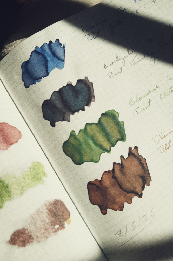 A vertical colour photograph of ink swatches on a notebook with a grid pattern on the paper. The sun falls across the paper with a diagonal streak of shadow on the top right hand corner. The inks are blue, grey, green, and brown.