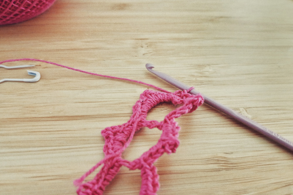 A horizontal colour photograph of a small lace crochet work in a pink cotton yarn on a desk. The hook is attached to the work and is also pink but a lighter shade.