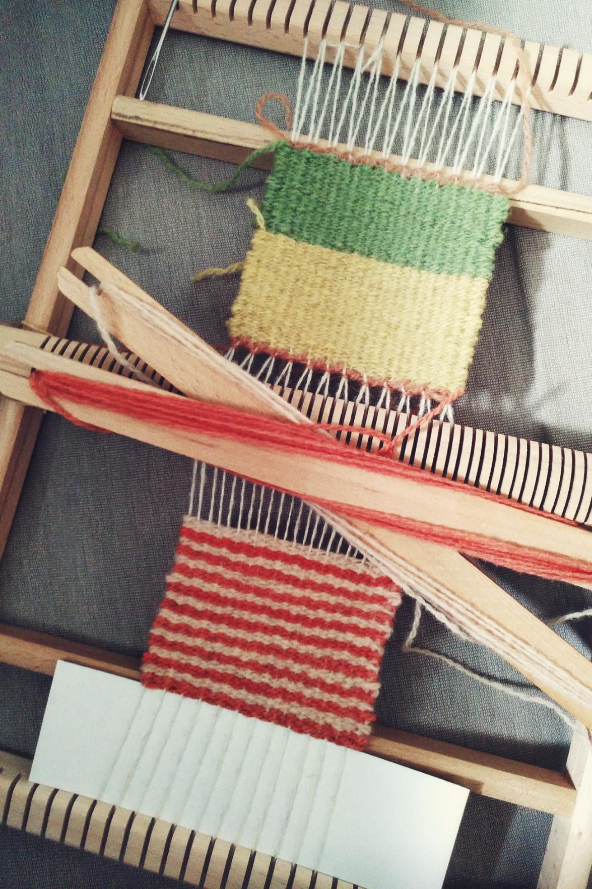 A vertical colour photograph of a small wooden loom on a grey sofa fabric with a finished yellow and green coaster on one side. On the other side is an in progress coaster in two shades of pink arranged in stripes.