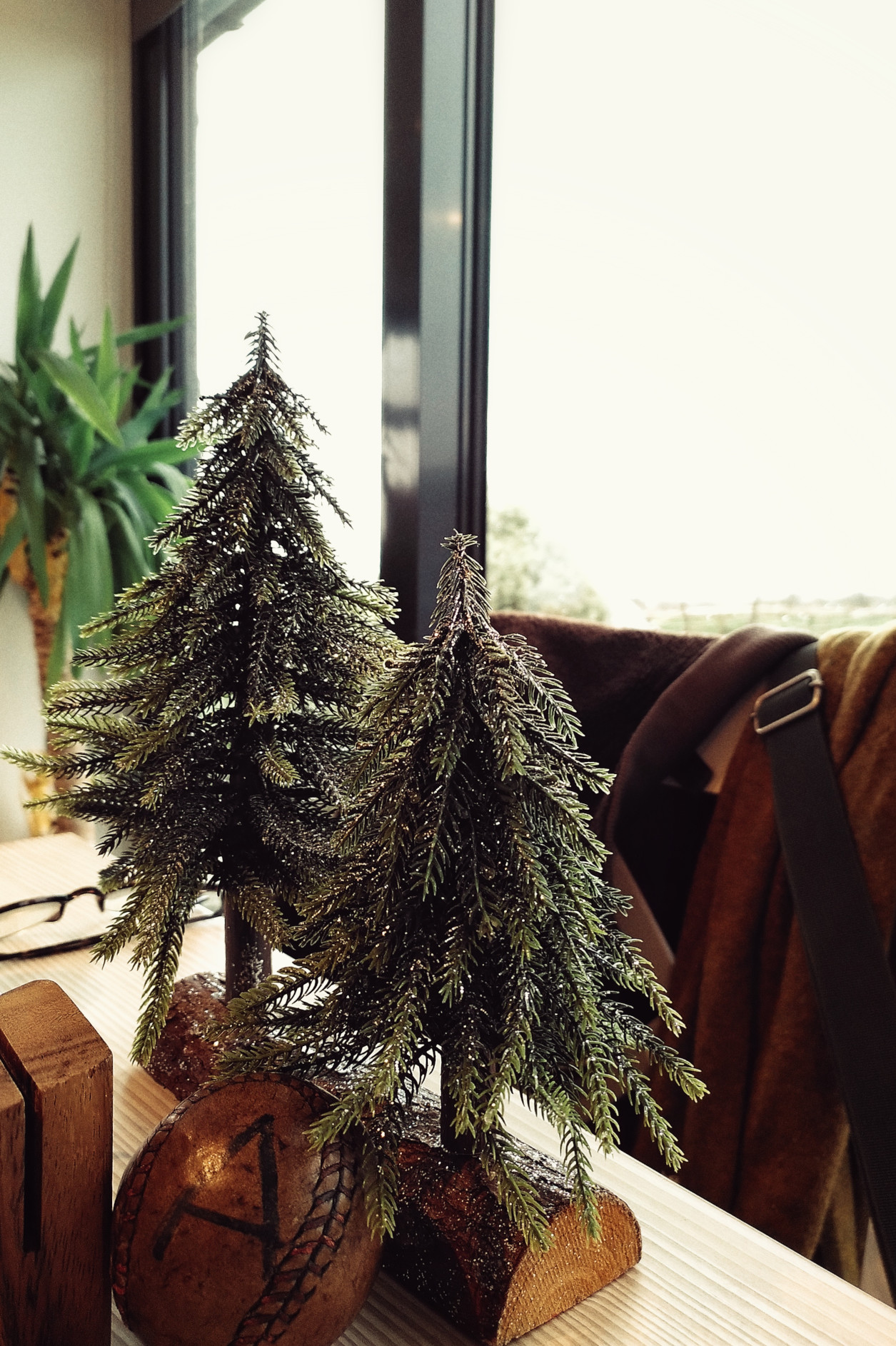 A vertical colour photograph of the edge of a restaurant table with two small plastic glittery Christmas tree planted on a log. In the background there is a chair with coats and bags.