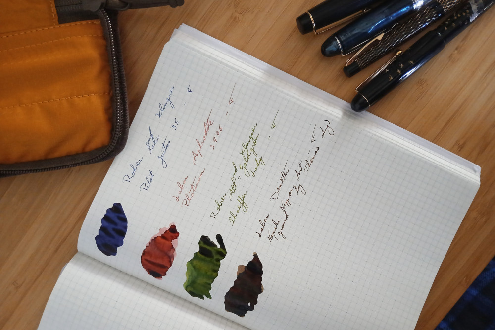 A horizontal colour photograph of ink swatches on a notebook with a grid pattern on the paper. The inks are blue, pink, green, and brown. Next to the notebooks are the pens and a pen pouch. Those are cut off by the frame.