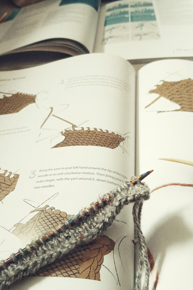 A vertical colour photograph of a small knitting work on colourful wooden needles. The yarn is grey. The work rests on a book opened on a page with purl stitch instructions.