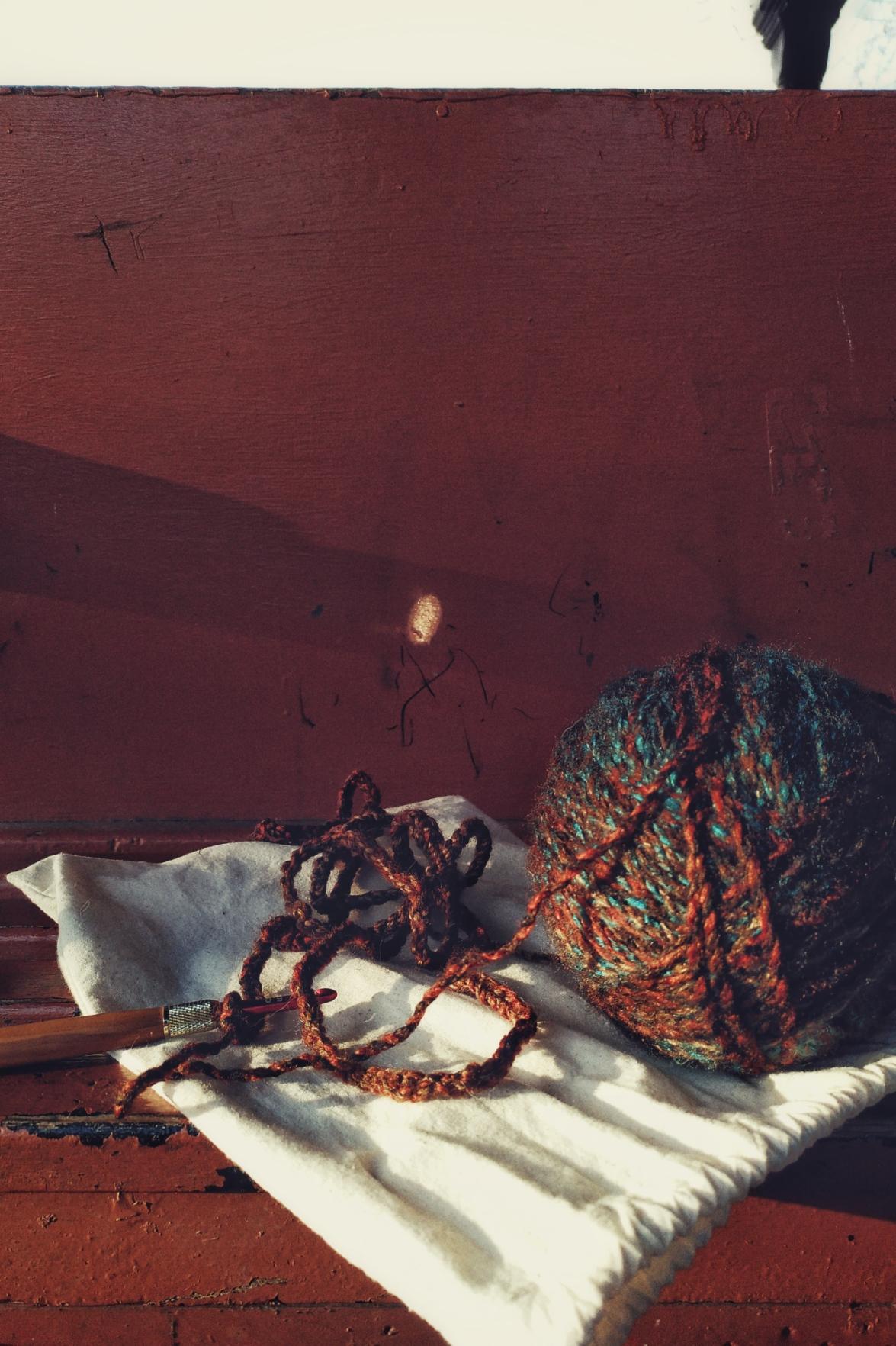 A vertical colour photograph of a long crochet chain with a ball of multicoloured yarn next to it. A red metal crochet hook with a wooden handle is at the end of the chain. The yarn is on a cream cloth. The whole is on a bench.
