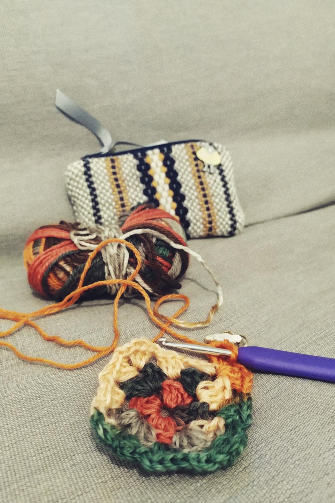 A vertical colour photograph of a colourful small granny square on a grey sofa. A crochet hook is still attached to the square. Behind is the yarn used. It's a colour changing yarn. Behind that is a woven yellow, cream, and black small pouch with a sheep pin in it.