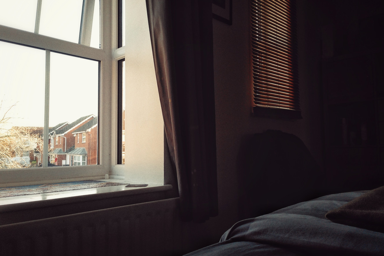 A horizontal colour photograph of a dark bed corner and a window full of light. Outside there is a row of brick houses and overblow bare tree branches.