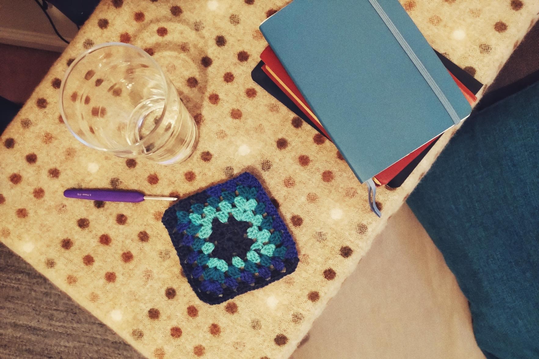 A horizontal colour photograph of a crochet granny square in a variegated blue yarn. Next to it is a metal crochet hook with a purple handle. They rest on a table covered with a polka dot brown woollen cloth. On the table is also a pint glass with some water and a pile of notebooks.