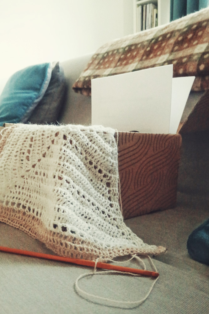 A vertical colour photograph of a crochet shawl in progress. The shawl is mostly white with two rows of creamish pink colour. It drapes over a carboard box on a grey sofa bed.