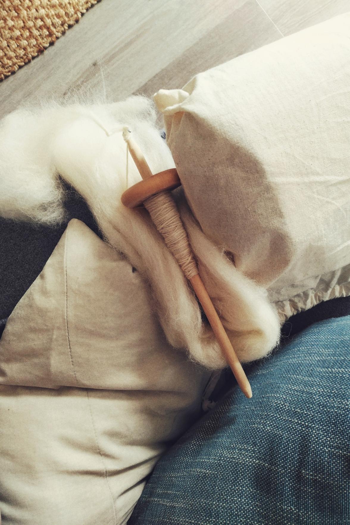 A vertical colour photograph of a wooden drop spindle with white spun wool on the shaft. It rests on a bit of white wool fibre but a white linen bag. The whole rests on a grey sofa with a white pillow and a turquoise pillow. The floor beyond the sofa is a light grey brown. In the top far left corner a bit of a jute mat is visible.