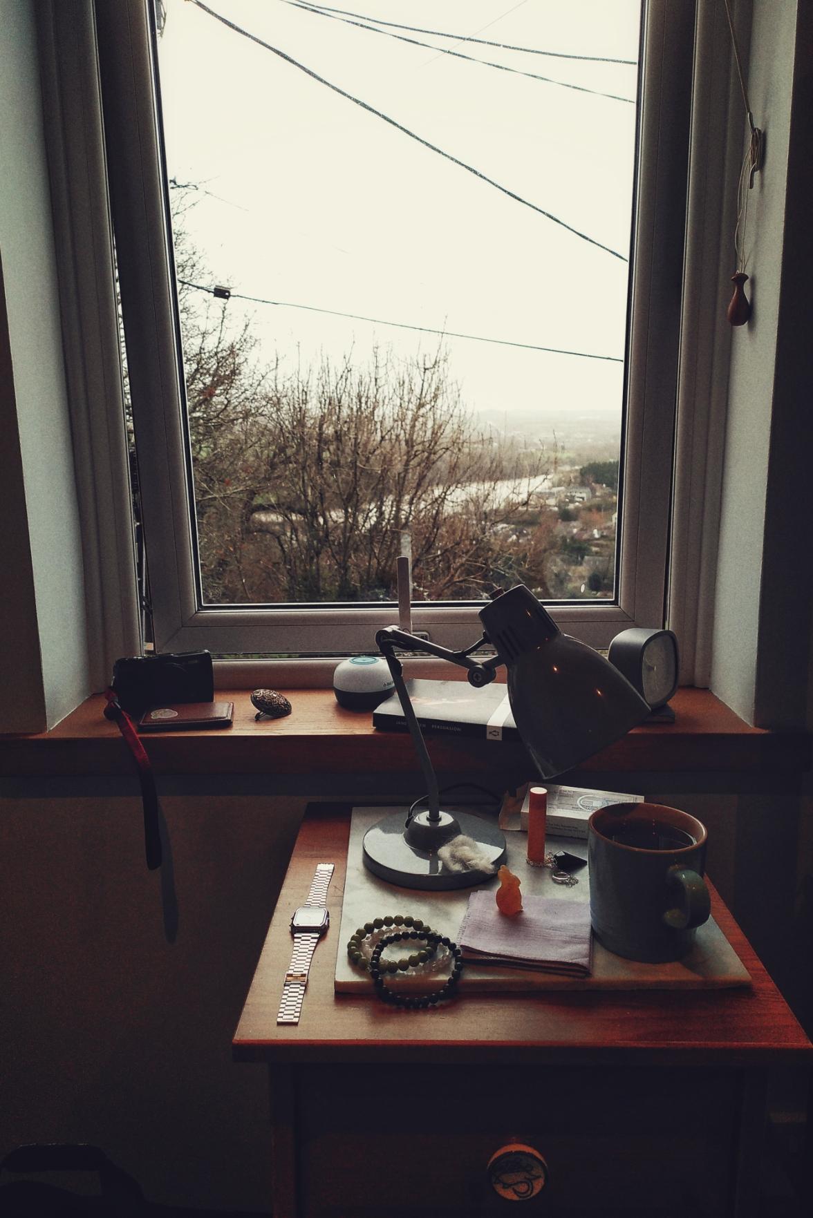 A vertical colour photograph of a bedside table with a grey lamp on it, as well as an assortment of bracelets, a watch, an amber rabbit, a handkerchief, a mug full of tea, and some wool. Behind is a window ledge with a camera, a bronze brooch, a book, a lamp, and a small clock. The window is opened a little at the bottom. Outside, the trees are bare, wires hang in the air, and a river is visible in the distance before clouds obscure the rest of the background.