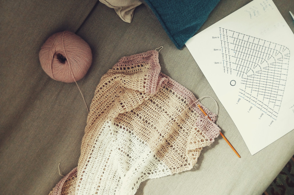 A horizontal colour photograph of a crochet shawl in progress on a grey sofa. The shawl has three colours on: white, cream, and pinkish violet. The hook is orange. On the other side, the ball of yarn rests. Next to the hook is a diagram of the work.