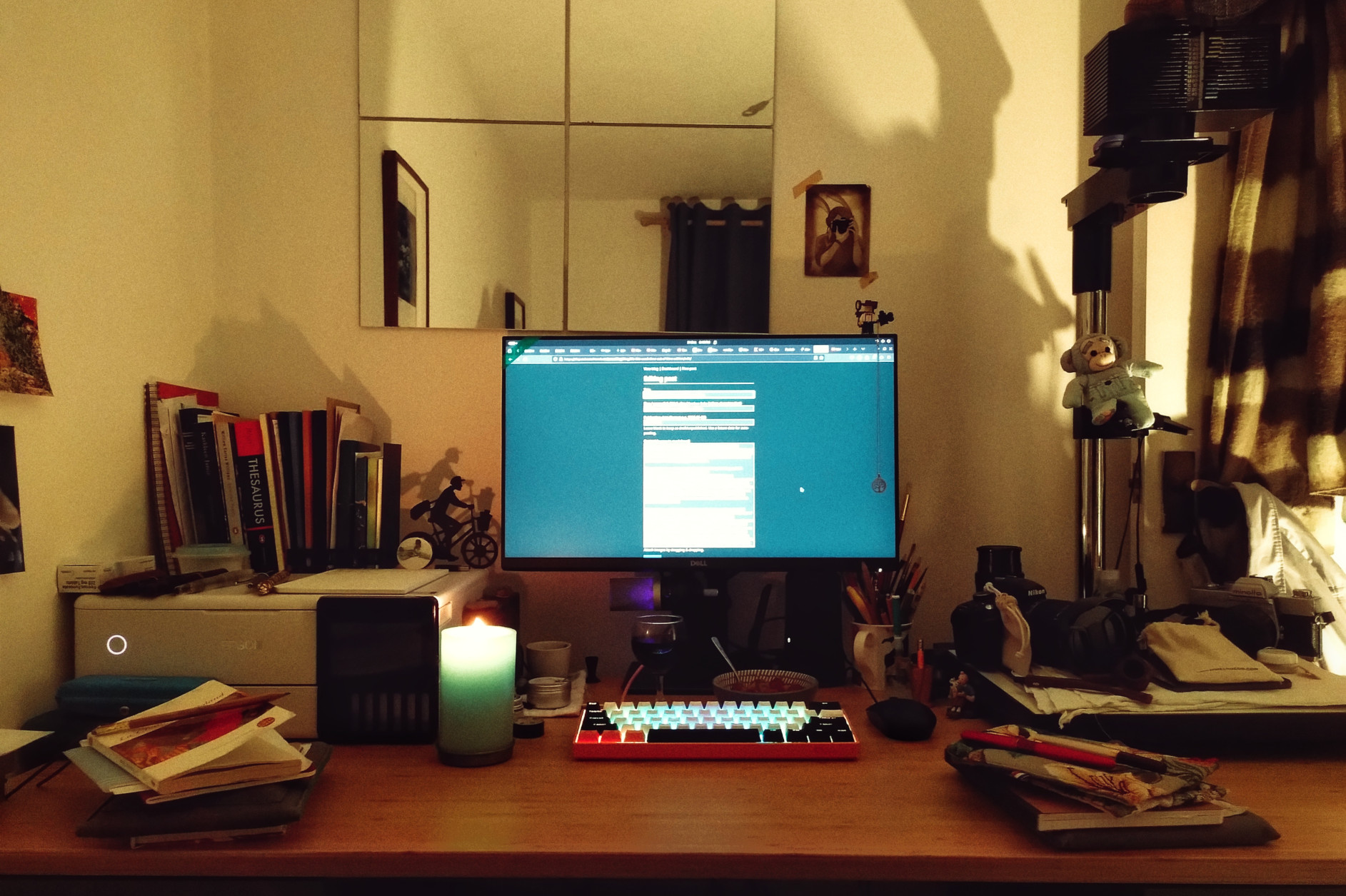 A horizontal colour photograph of a desk with a computer screen, keyboard, candle, papers, books, pens, printer, enlarger, and other paraphernalia. The computer screen is lit up to a writing program though none of the words are visible.