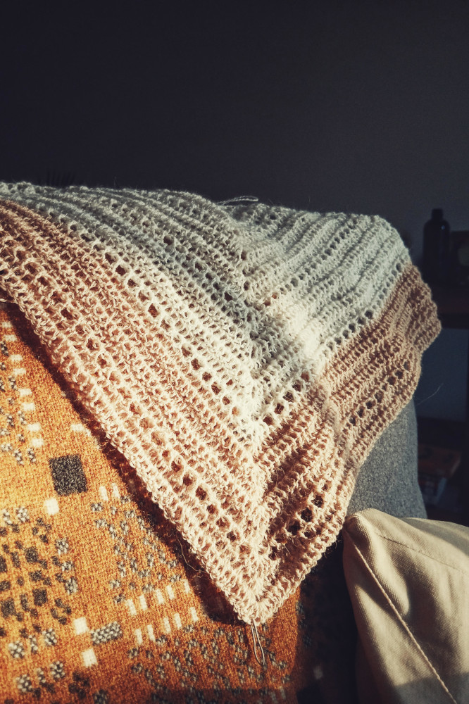 A vertical colour photograph of a crochet shawl in progress. A part of it is white, another creamish pink. It rests on a yellow and grey Welsh woven blanket.